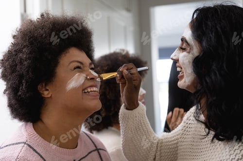 Preview: Happy biracial young woman with beauty cream on face applying face pack to female friend at home