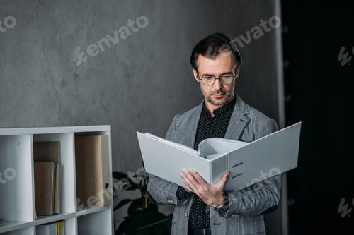 Preview: serious businessman in eyeglasses holding folder and working with papers in office