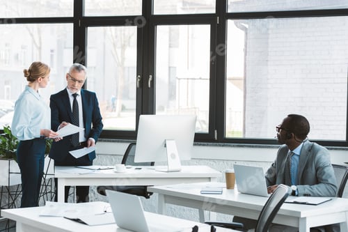Preview: african american businessman using laptop and looking at colleagues discussing papers in office