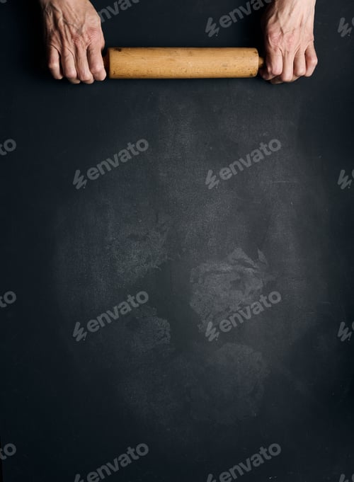 Preview: Two female hands hold a wooden rolling pin for rolling dough on a black table, top view.