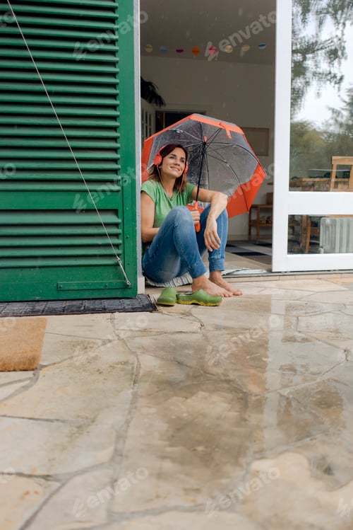 Preview: Smiling mature woman sitting on terrace in rain under umbrella listening to music with headphones