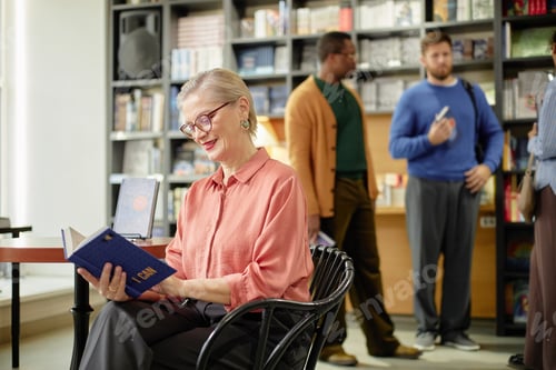 Preview: Senior Caucasian Woman Reading Book in Library with Diverse People Socializing