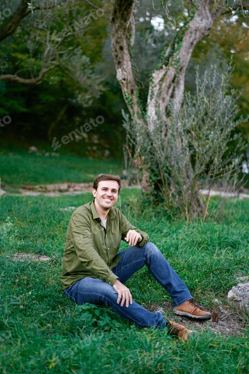 Preview: Young man sitting on green grass near the tree