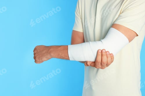Preview: Man with arm wrapped in medical bandage on light blue background, closeup