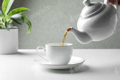 Preview: Pouring freshly brewed tea from teapot into cup at white table, closeup