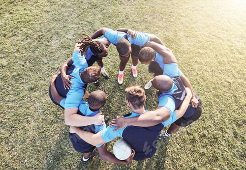 Preview: Above diverse group of rugby players standing in a huddle together outside on a field. Young male a
