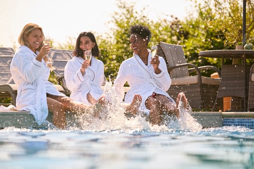 Preview: Group Of Mature Female Friends Wearing Robes Sitting With Feet In Pool Drinking Champagne On Spa Day