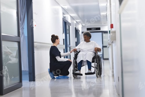Preview: Diverse female doctor talking with senior female patient in wheelchair in hospital corridor