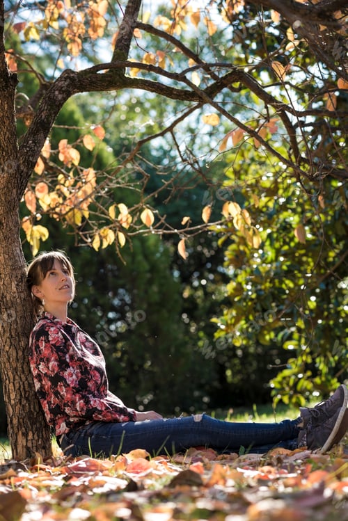 Preview: Young woman sitting under autumn tree leaning on its trunk