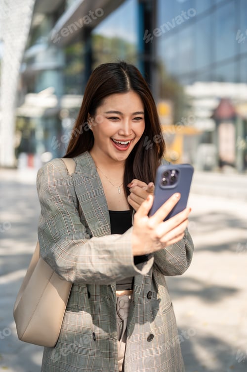 Preview: Pretty asian woman looking and pointing at smartphone in surprised while standing outside a building