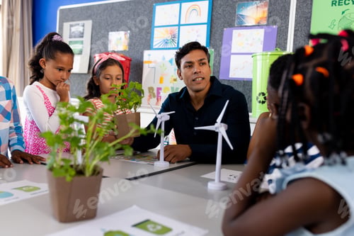 Preview: Happy diverse male teacher and children with windmills and plants in class at elementary school