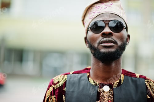 Preview: African stylish and handsome man in traditional outfit and cap standing outdoor.