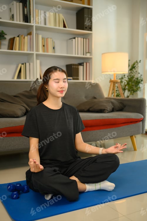 Preview: Young woman with eyes closed practicing meditation on a blue exercise mat in living room