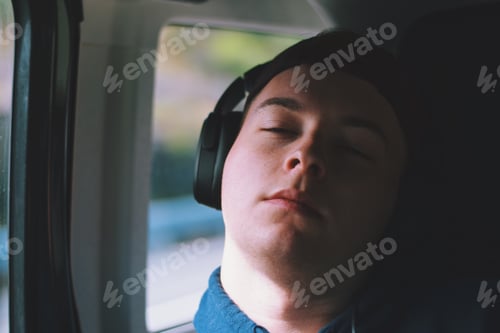 Preview: young man wearing headphones listening to music while riding in a car on a road trip sleeping