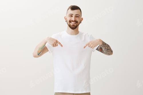 Preview: Look at my t-shirt. Positive carefree male friend with beard and tattoos on arms, pointing at chest