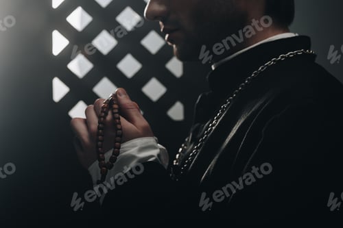 Preview: partial view of catholic priest holding wooden rosary beads near confessional grille in dark with