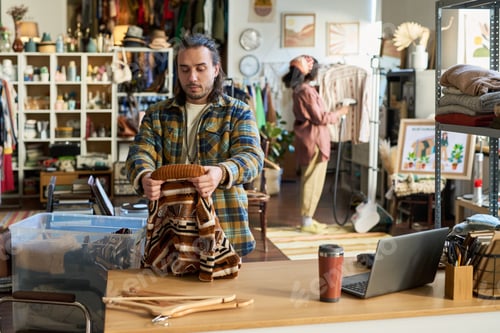 Preview: Young Adult Caucasian Man Sorting Clothing at Thrift Shop Checkout Counter