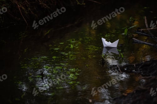 Preview: Simple, white, paper boat floats along small river in green forest. Hobby origami.