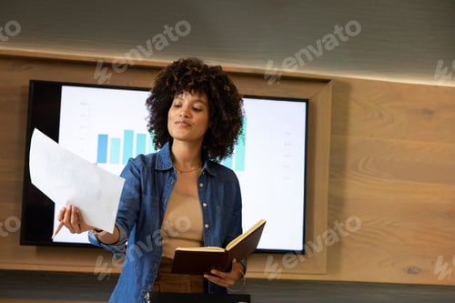 Preview: Adult female presenter in denim shirt handing paper holding notes at podium by bar chart monitor