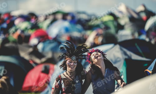 Preview: Two smiling young women at a summer music festival face painted, wearing feather headdress, standing