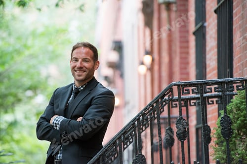 Preview: Business man in the city in a suit on the steps of a brownstone building. Arms folded.