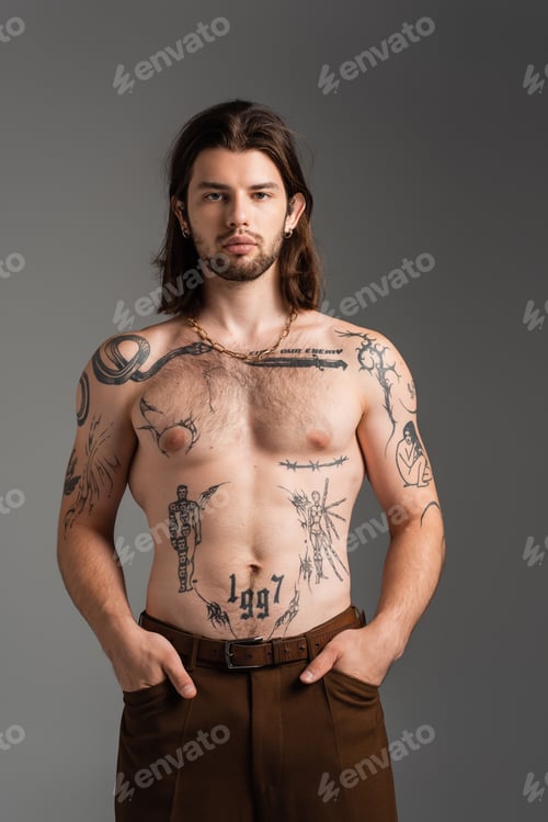 Preview: Long haired and tattooed man in pants posing isolated on grey