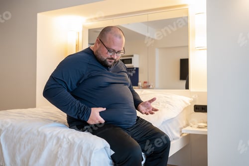 Preview: Huge man suffering from extra weight touching stomach sitting on bed in bedroom.