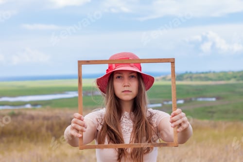 Preview: Girl in pink hat holds wooden picture frame on nature landscape background portrait Creative idea