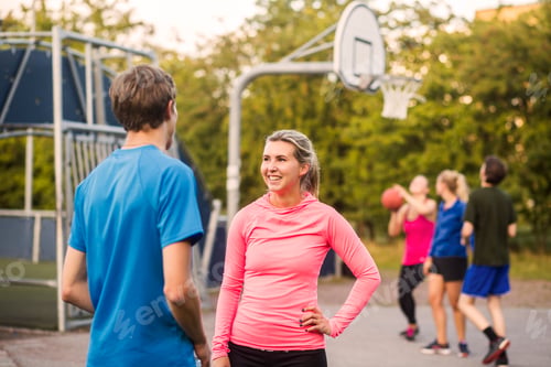 Preview: Happy woman talking to man while friends playing basketball at park