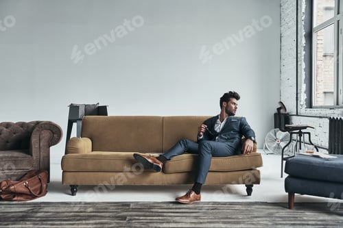 Preview: Enjoying free time. Thoughtful young man in full suit looking away while sitting on the sofa at home