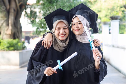 Preview: Two young Asian Muslim woman in graduation costumes look at camera, smiling and hugging.