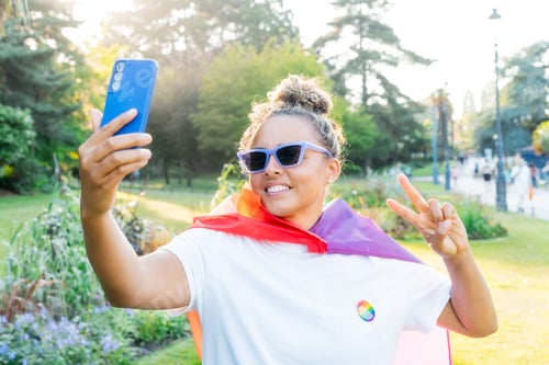 Preview: Young Latin happy woman wrapped in a rainbow flag, symbol of LGBT community, makes selfie on phone