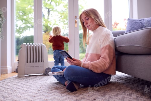 Preview: Mother With Son Trying To Keep Warm By Radiator At Home During Cost Of Living Energy Crisis