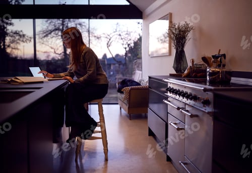 Preview: Evening Shot Of Woman In Kitchen Working Or Studying From Home Using Digital Tablet