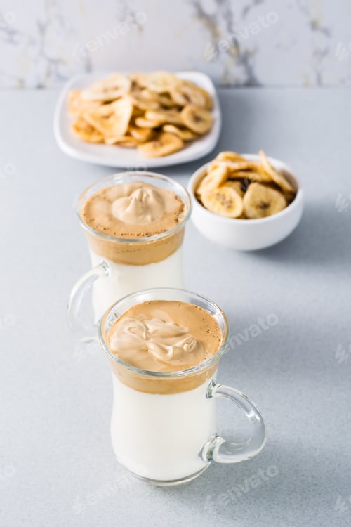 Preview: Two cups with dalgona coffee and banana chips on a gray background. Vertical view