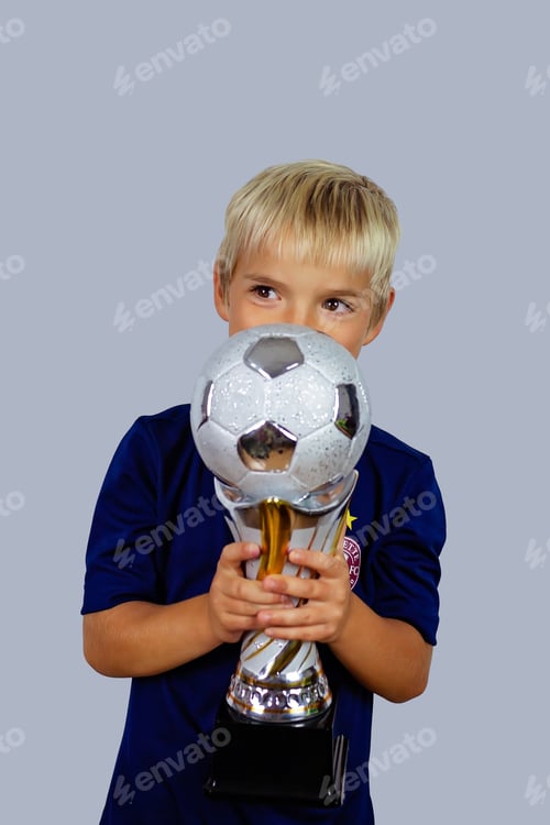 Preview: Young soccer player in blue jersey holds winners cup after the goal, isolated at light background