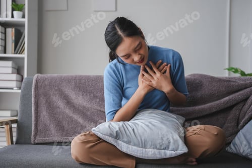 Preview: Young woman is sitting on her sofa, holding a pillow and experiencing discomfort in her chest