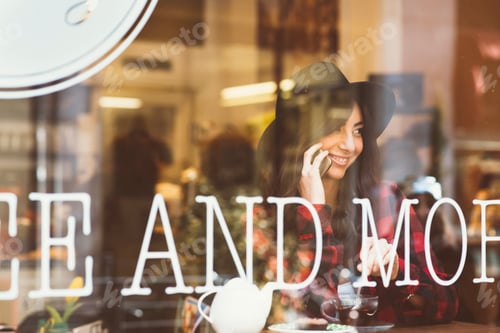 Preview: Young woman sitting in cafe, using smartphone, view through window