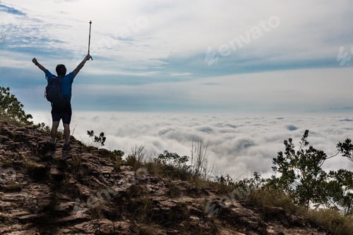 Preview: Hiker Asian man in silhouette standing on peak of mountain with cloud scape raises hand to signify