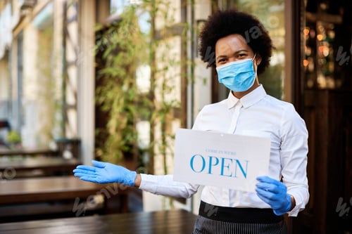 Preview: Black waitress holding open sign while wearing protective face mask and gloves at sidewalk cafe.
