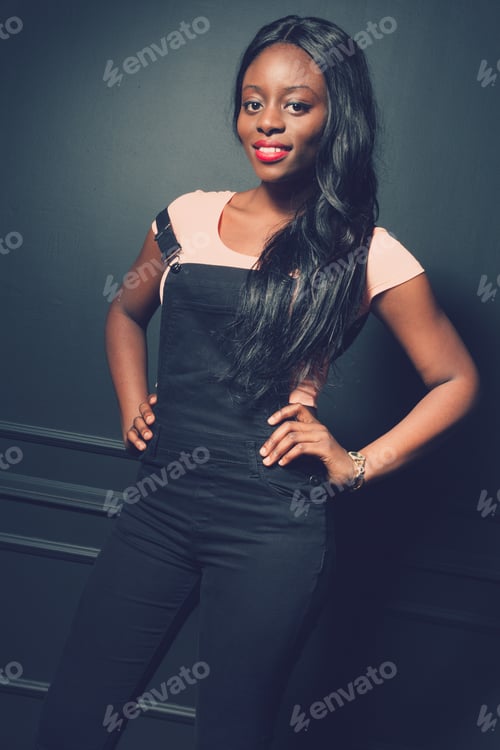Preview: Young african woman posing in overalls against a dark gray wall