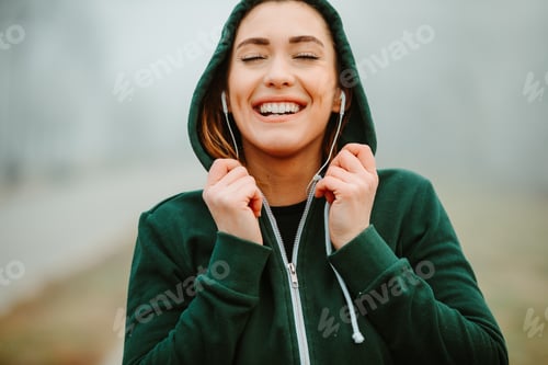 Preview: Close photo of beautiful sportswoman with hoodie and headphones during outside training session.
