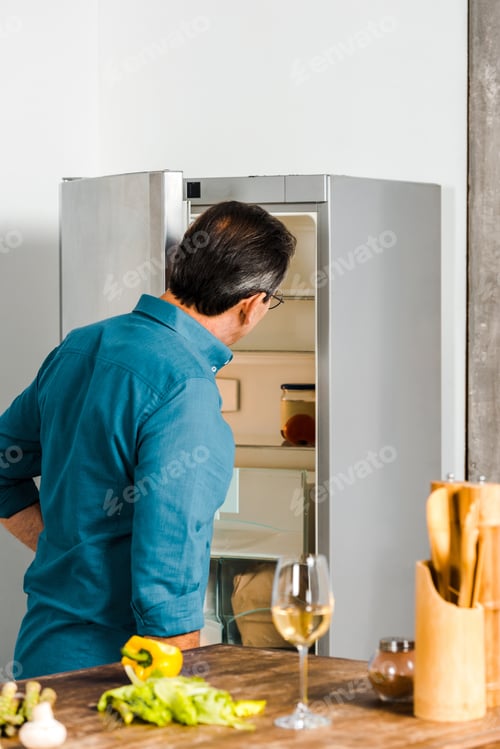 Preview: back view of mature man looking into open fridge in kitchen