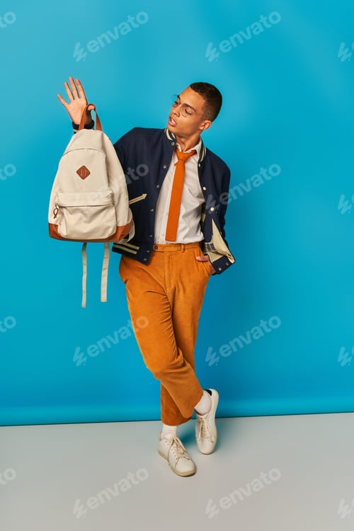 Preview: african american student in orange pants standing with hand in pocket and backpack on blue