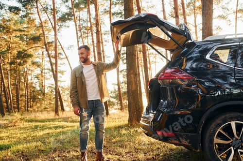 Preview: Near the car. Man in jeans is outdoors in the forest with his black colored automobile