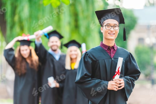 Preview: selective focus of smiling asian graduate with classmates behind in summer park