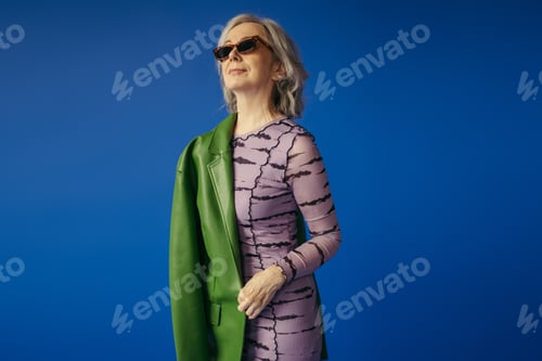 Preview: senior woman in sunglasses and dress standing with green leather jacket isolated on blue