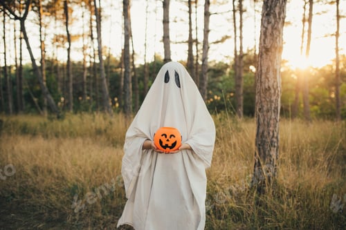 Preview: Girl wearing ghost costume holding pumpkin bucket with candies, standing in a forest.