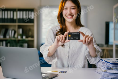 Preview: Asian accountant woman is smiling while sitting at her desk and holding up a credit card. She is sur