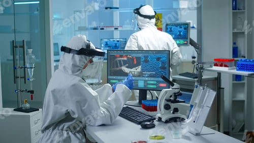 Preview: Chemist in ppe suit working at pc while lab technician bringing her blood samples